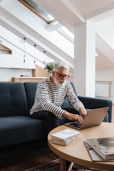 Elderly man with grey hair and glasses using a laptop on a sofa in a bright, modern room.