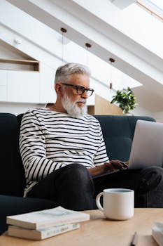 Elderly man with eyeglasses working on a laptop in a stylish room. Coffee and books on the table.