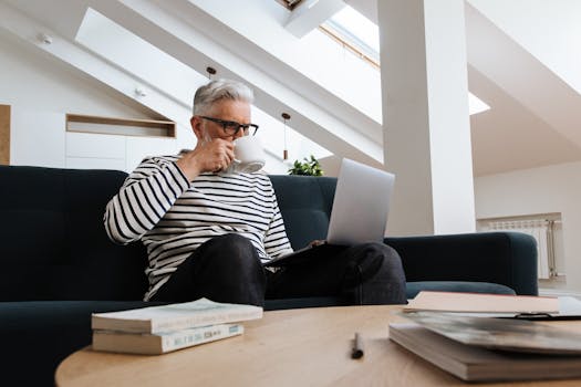 A senior man enjoying coffee while working on a laptop from home in a modern indoor setting.