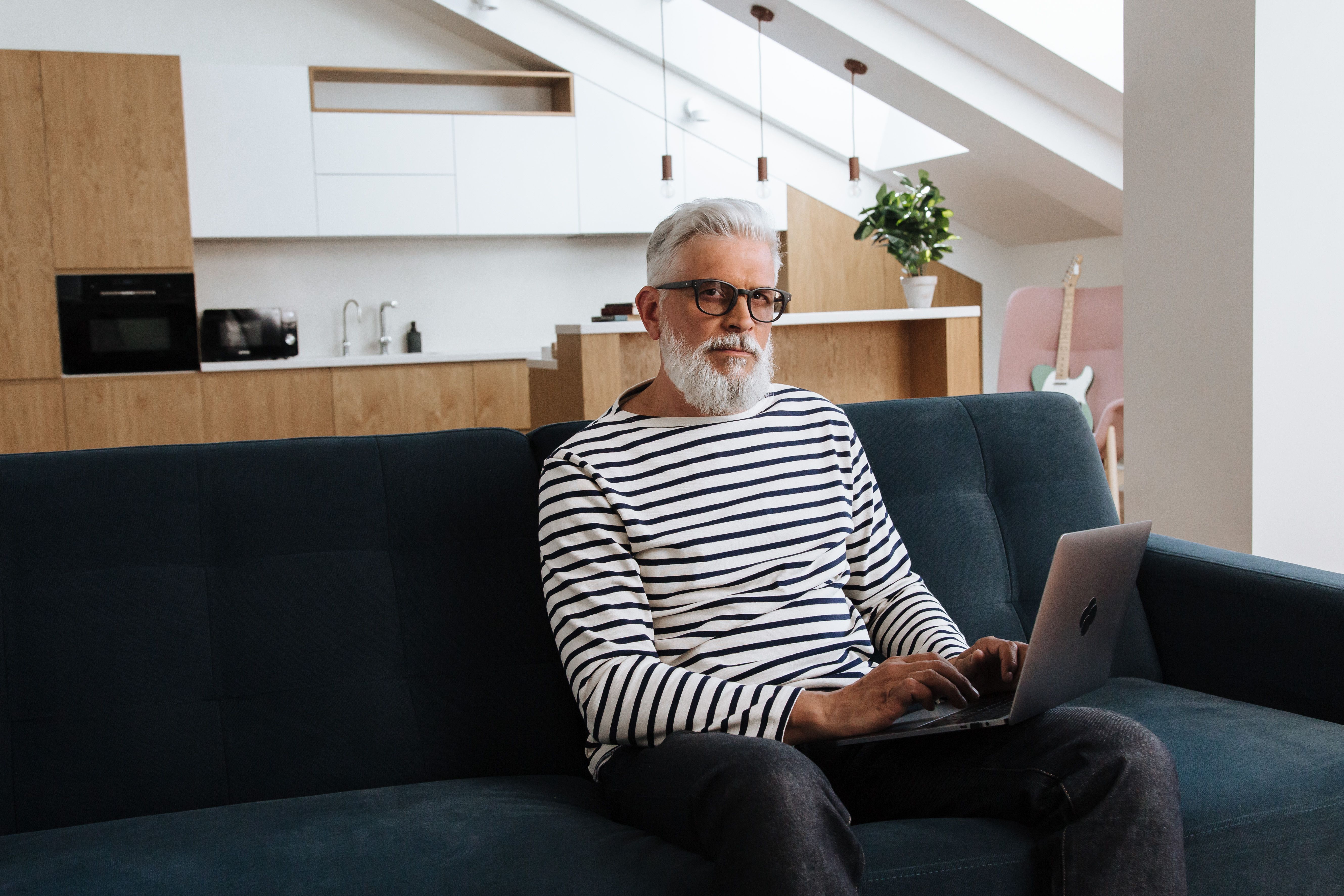 Elderly man with gray hair and beard using laptop on couch in stylish apartment.