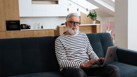Elderly man with gray hair and beard using laptop on couch in stylish apartment.