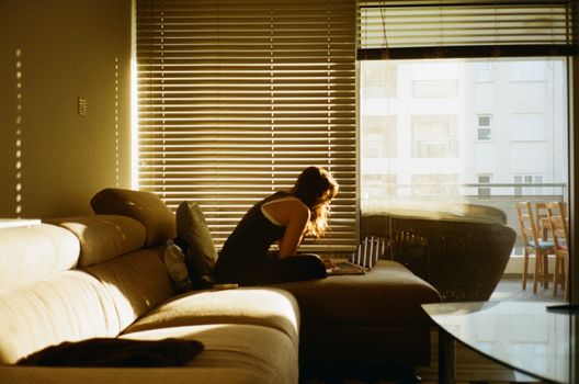 A woman sits on a sofa using a laptop near a window with blinds in Limassol, Cyprus.