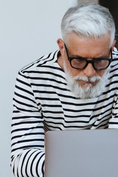 Senior man with grey beard and glasses focused on laptop work at home.