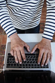 Overhead shot of a man in a striped shirt typing on a laptop indoors.