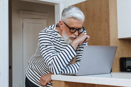 A senior adult man with glasses concentrating on his laptop at home, symbolizing modern working environments.