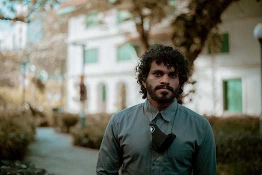 A contemplative man with curly hair and a beard posing outside in a serene setting.