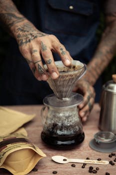 Tattooed barista's hands brewing coffee using a pour-over method at a café setting.