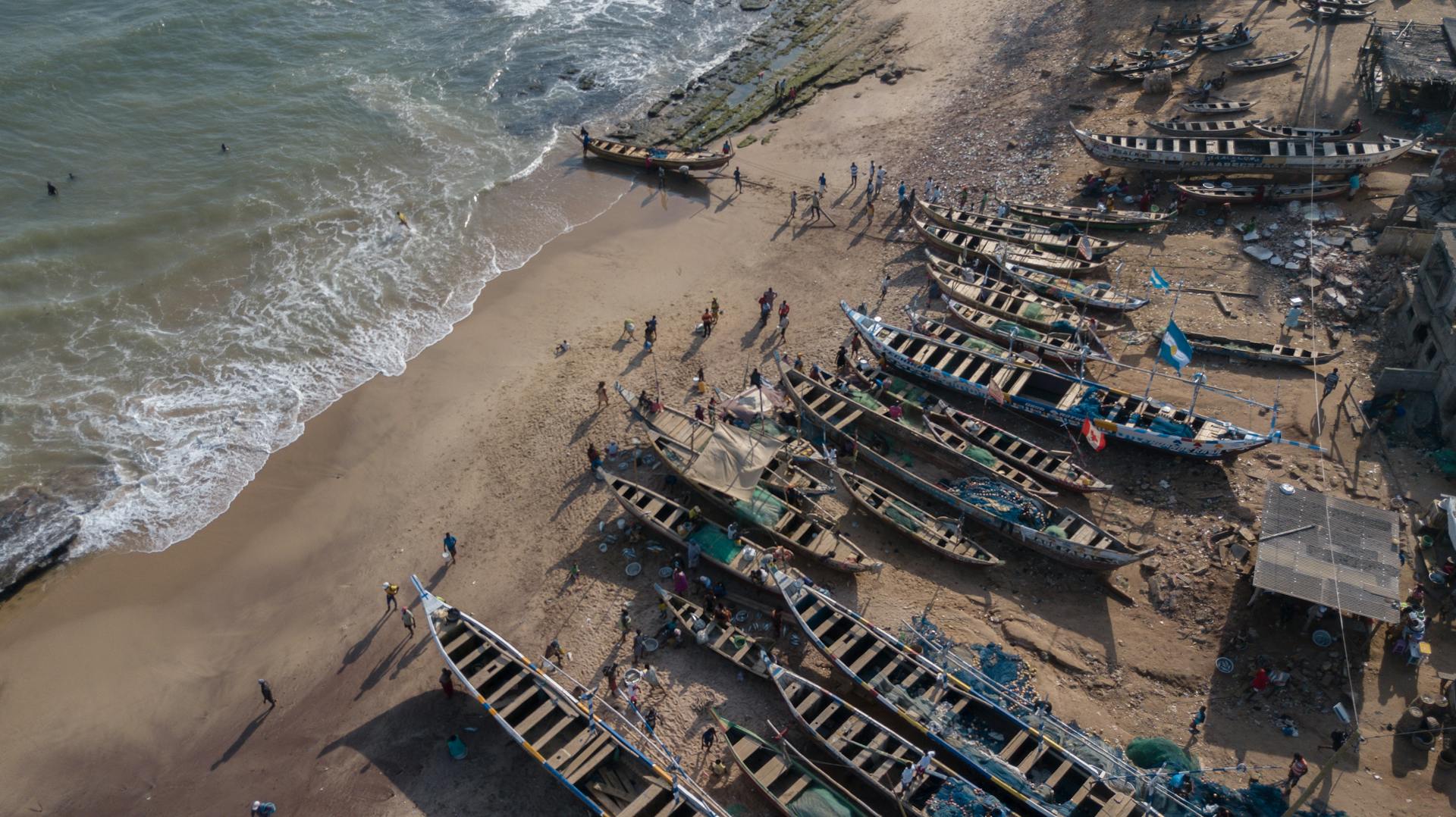 Aerial view of colorful fishing boats docked at Prampram beach in Ghana with people on the sandy shore.