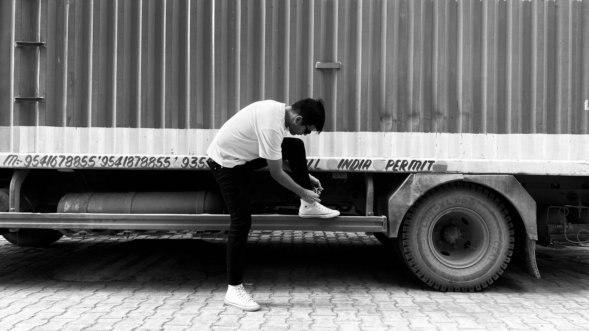 A stylish monochrome photo of a man tying shoelaces on the step of a lorry in India.