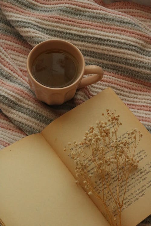 A Mug with Hot Drink Beside an Open Book with White Flowers