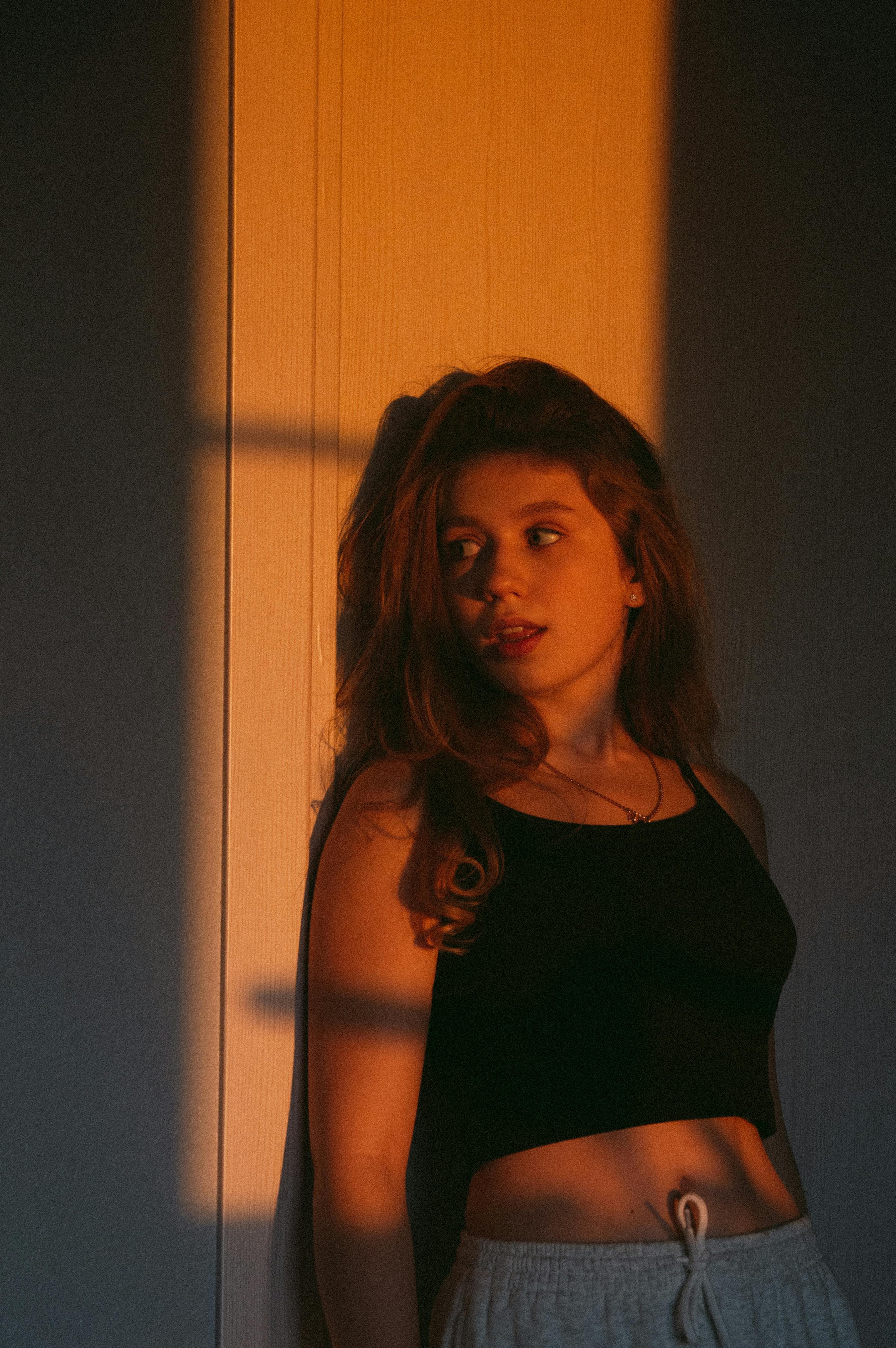 Young woman in a dark room, standing against a wall with warm sunlight casting shadows.