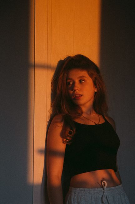 Young woman in a dark room, standing against a wall with warm sunlight casting shadows.
