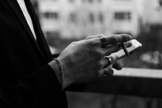Close-up of a hand holding a smartphone and cigarette with rings and bracelet in grayscale.