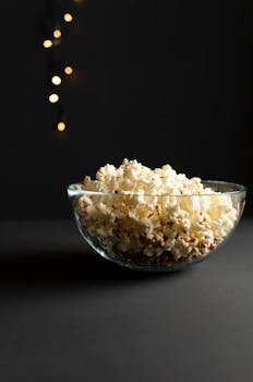 Glass bowl filled with fresh popcorn against a dark backdrop with soft bokeh lights, creating a cozy ambiance.
