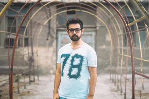 Free Young bearded man in eyeglasses stands confidently under an urban metal archway, wearing a stylish number 78 t-shirt. Stock Photo