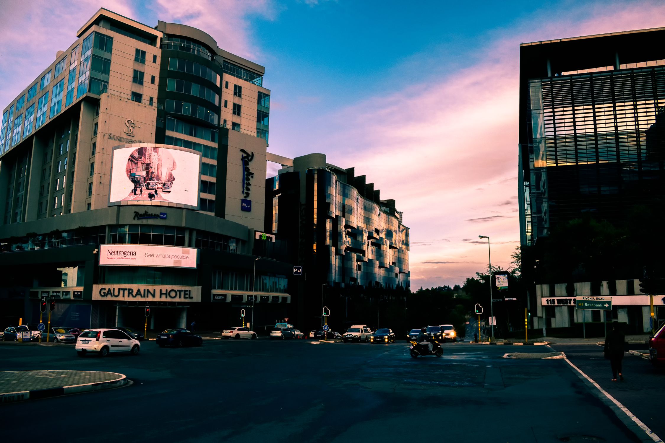 A bustling city street with modern skyscrapers and vehicles at sunset, Gautrain Hotel visible