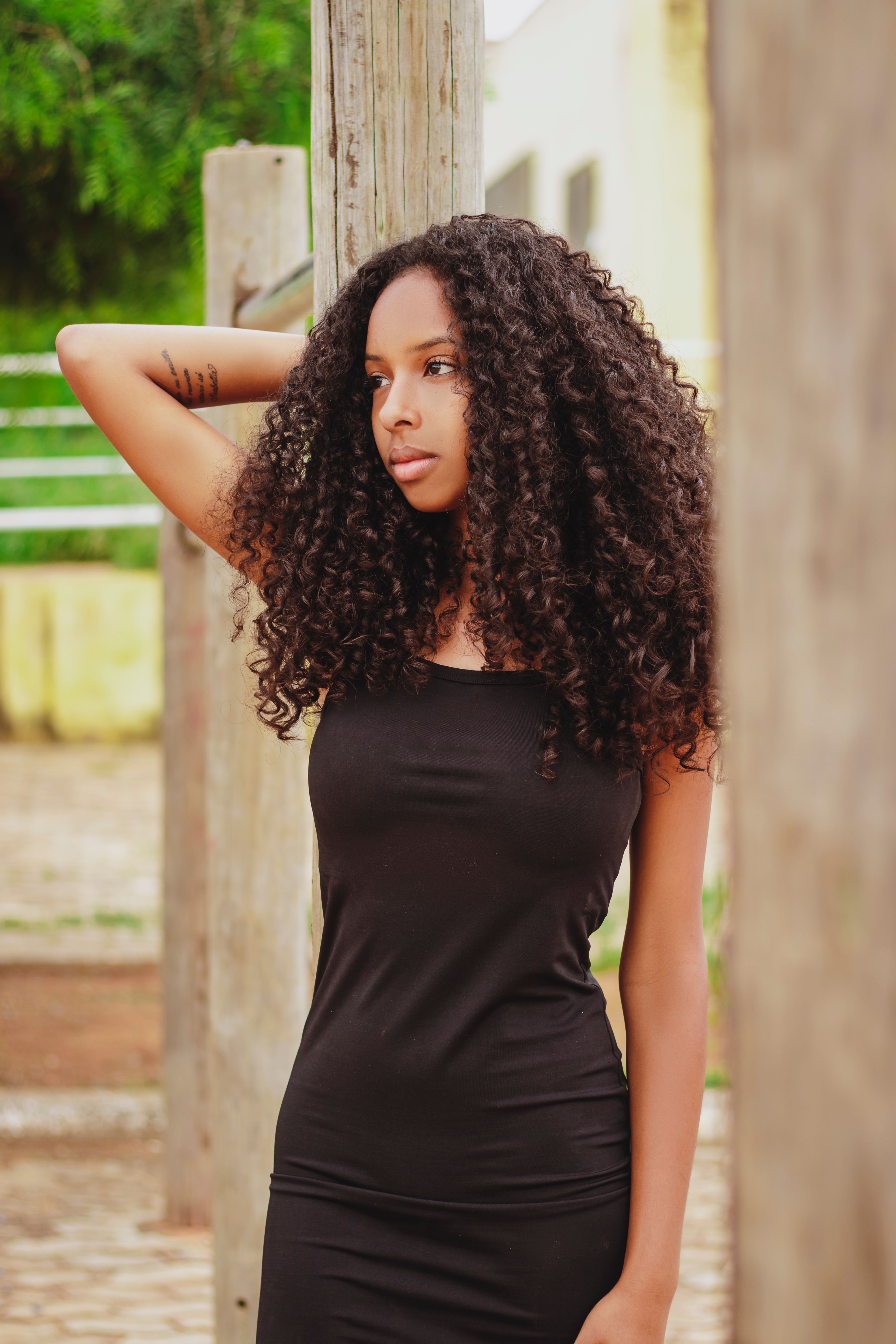 Beautiful outdoor portrait of a woman in a black dress with curly hair standing by wooden posts.