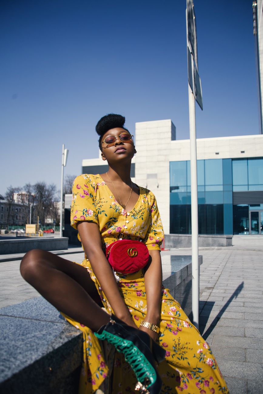 Mulher elegante em um vestido amarelo florido posando ao ar livre em um cenário urbano.