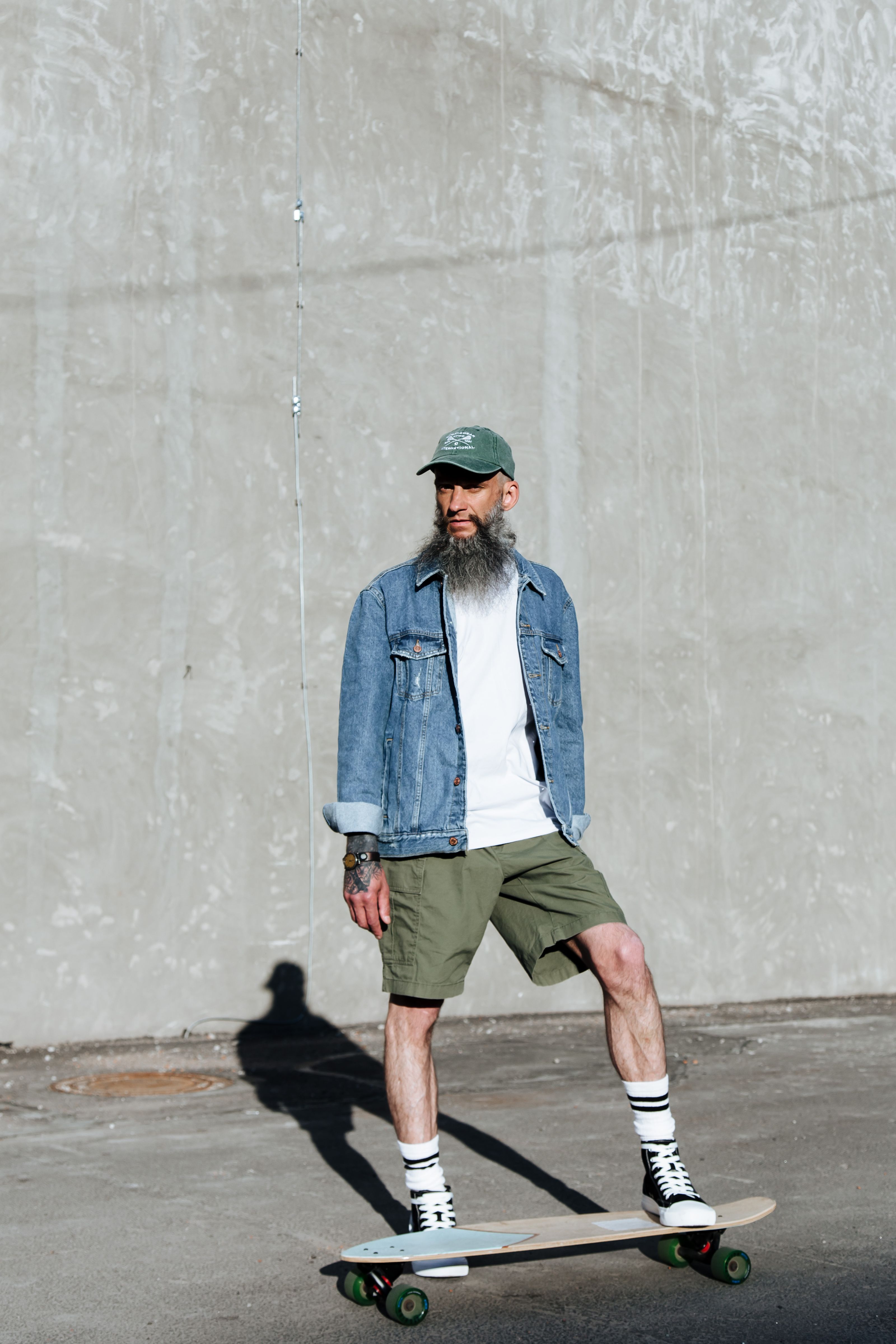 Homem fashion com barba andando de skate contra uma parede de concreto durante o dia.