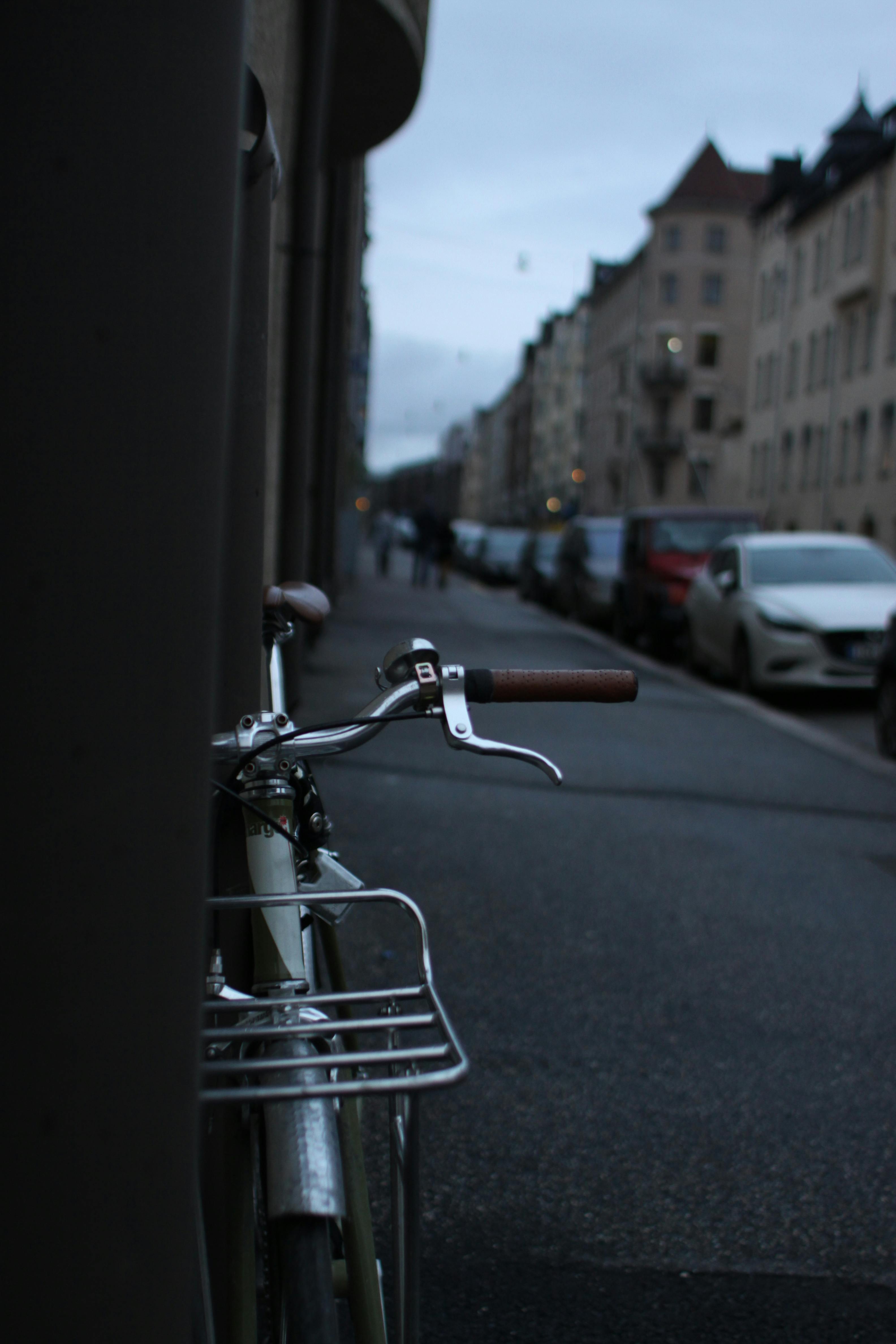 A bicycle is parked along a quiet urban street with parked cars and buildings in the background during dusk.