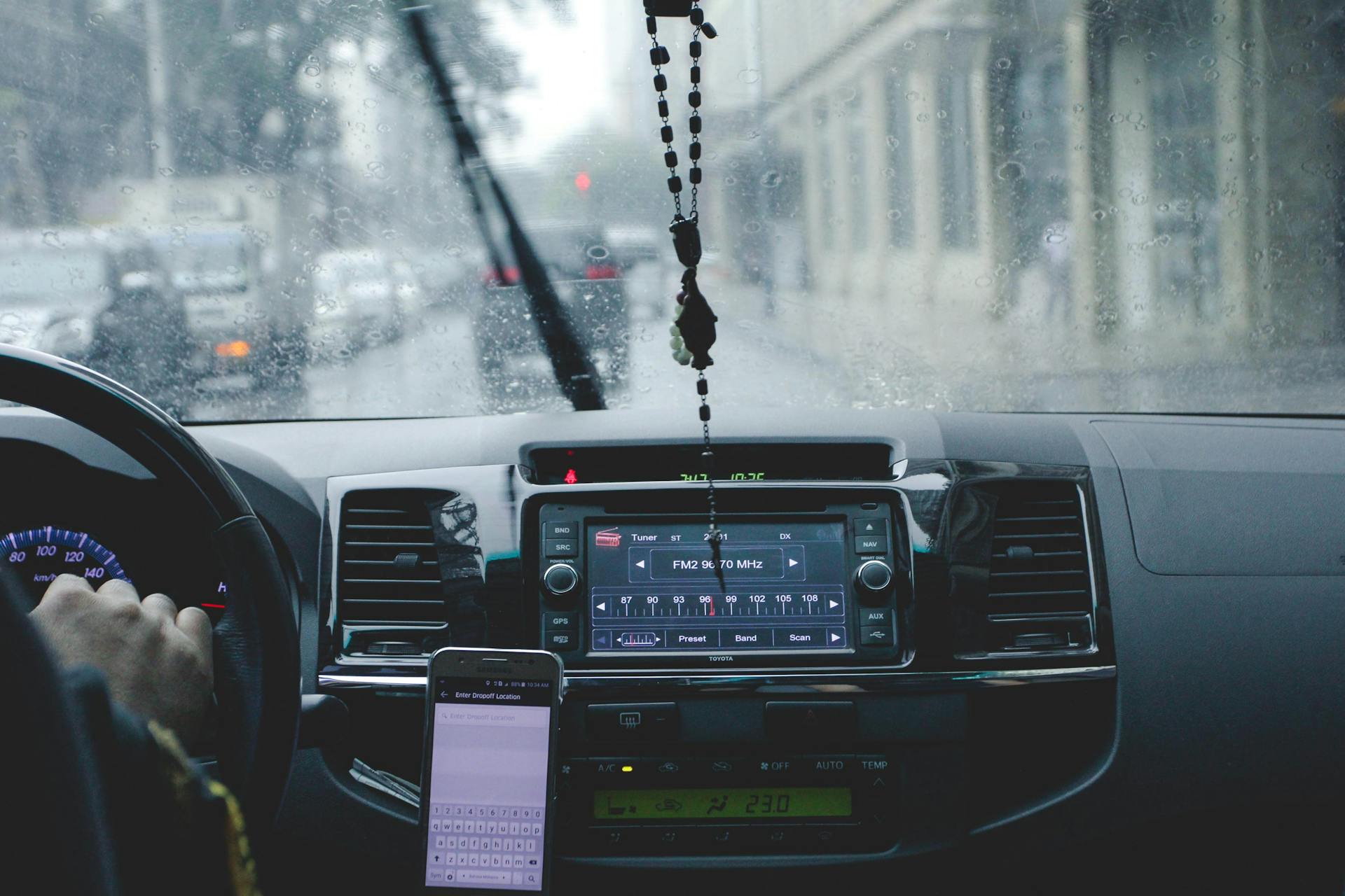 Car interior with driver navigating rainy cityscape using phone and dashboard.