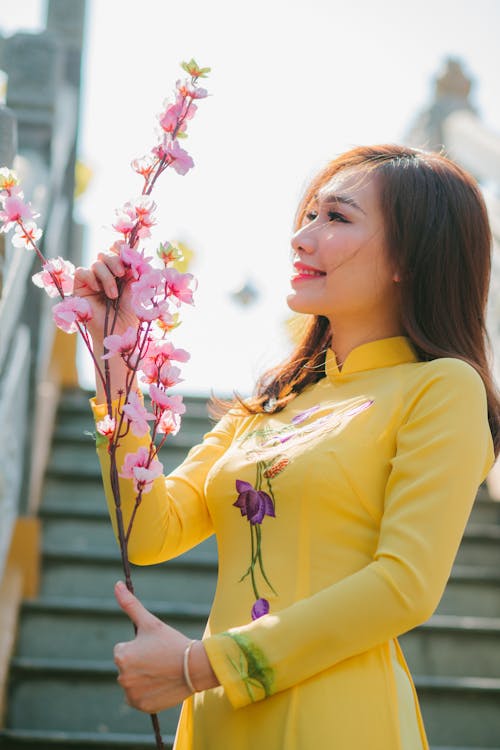Smiling Woman in Yellow Floral Dress Holding Delicate Pink Flowers