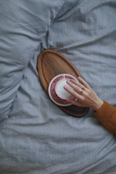 A hand holds a coffee cup on a wooden tray resting on a bed, creating a warm, cozy ambiance.