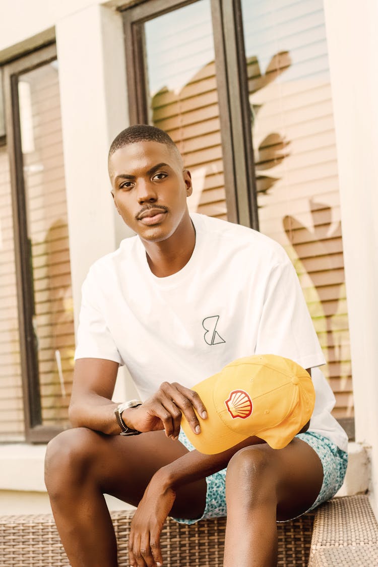 Man In White Shirt Sitting By Windowsill Holding Baseball Hat