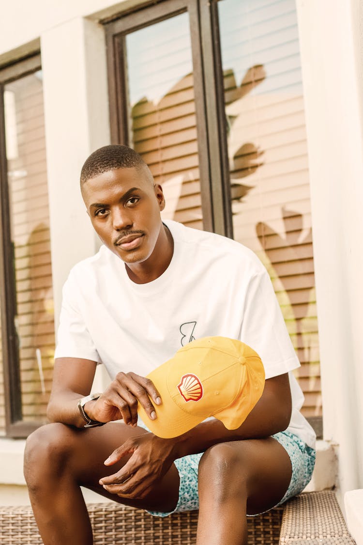 Man In White Shirt Sitting By Windowsill Holding Baseball Hat