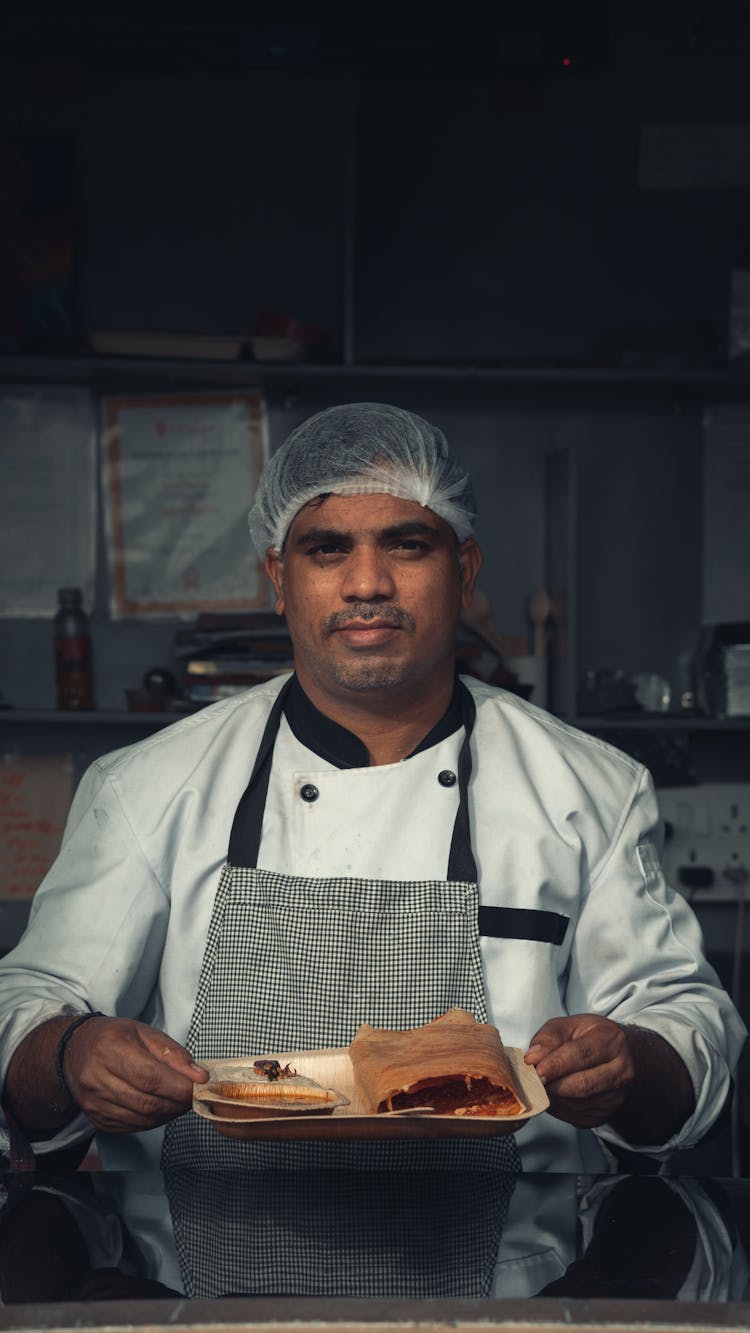 Man In White Chef Uniform Holding A Plate Of Food While Looking At The Camera