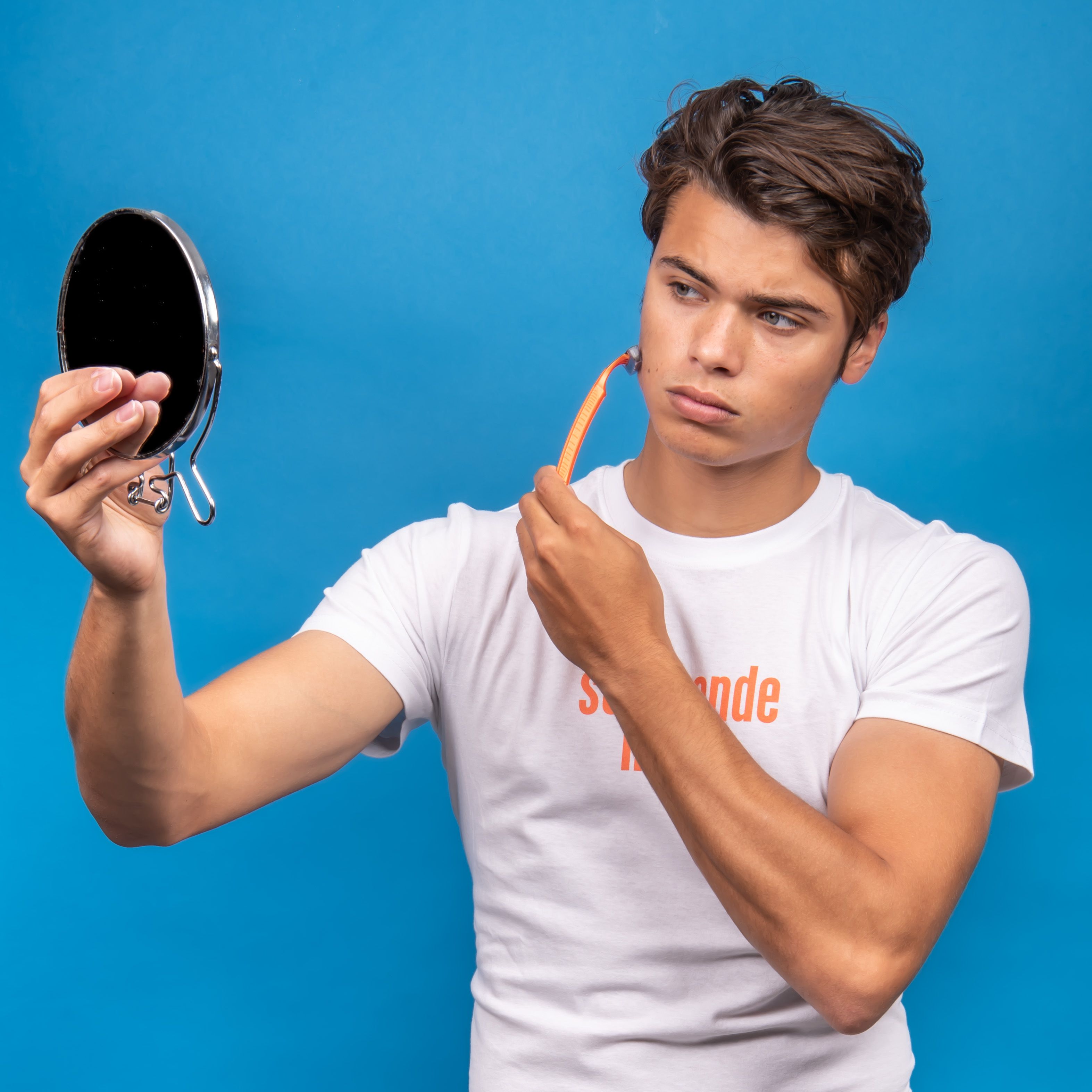 Young man in white shirt shaving and holding a mirror against a blue background.