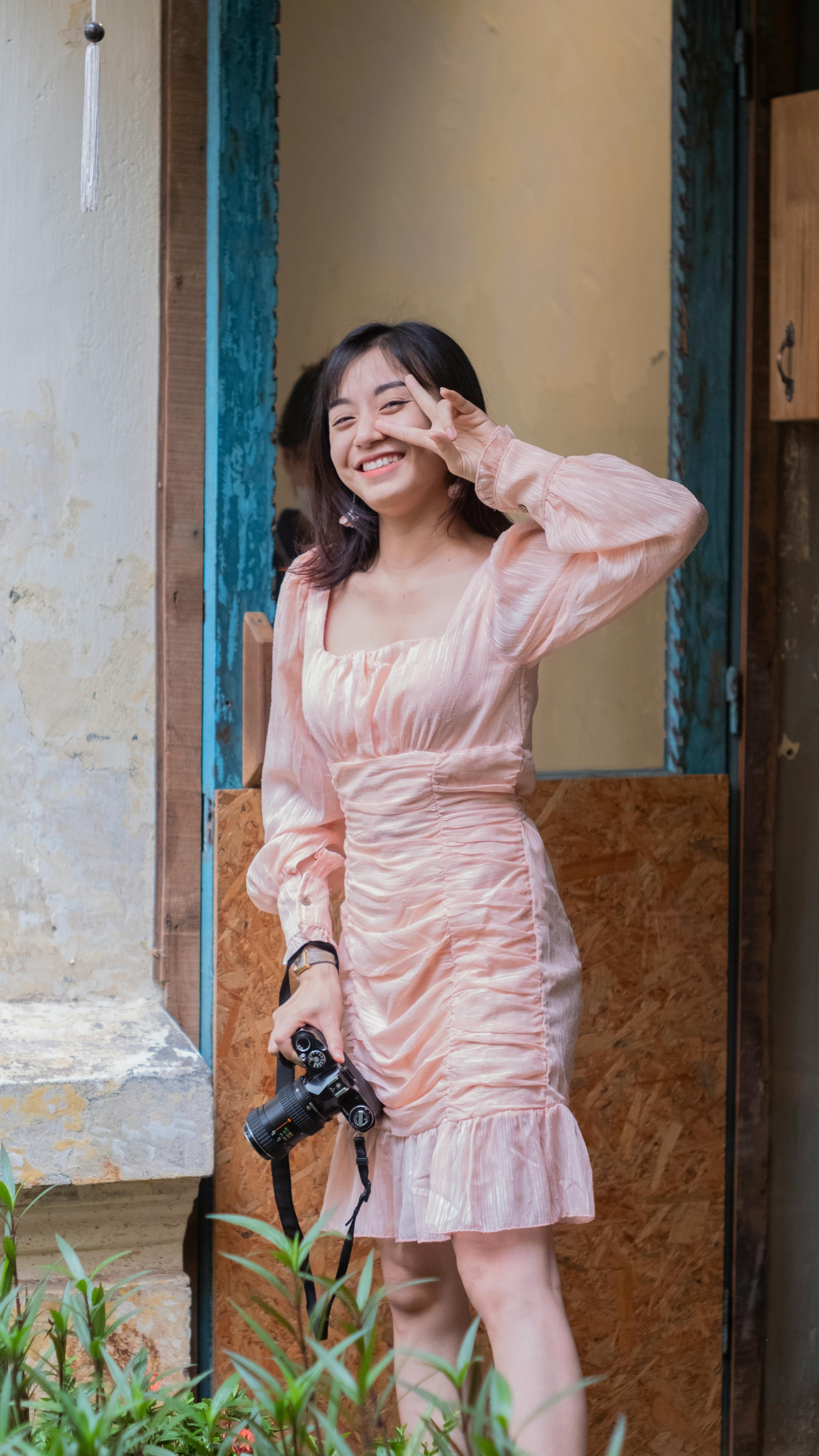 Joyous young woman in a pink dress holding a camera and making a peace sign indoors.