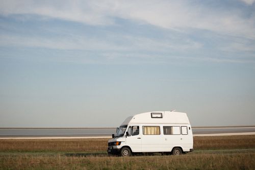 Free A white camper van parked on a grassy field by a tranquil shore under a clear sky. Stock Photo