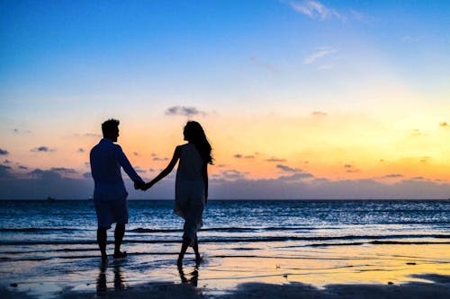 無料 日の出の間に海岸を歩く手をつないでいる男と女 写真素材