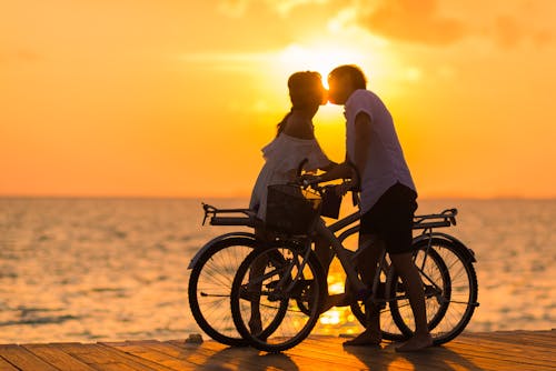 Photography of Man Wearing White T-shirt Kissing a Woman While Holding Bicycle on River Dock during Sunset