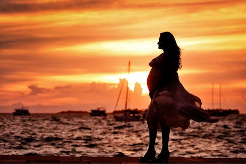 免費 孕婦在日落時站在海邊附近 圖庫相片