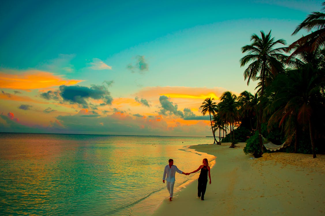 Man and Woman Holding Hand Walking Beside Body of Water during Sunset