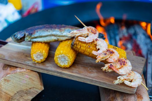 Close-up of grilled prawns, fish, and corn over open flame on a cutting board, perfect for outdoor barbecue scenes.