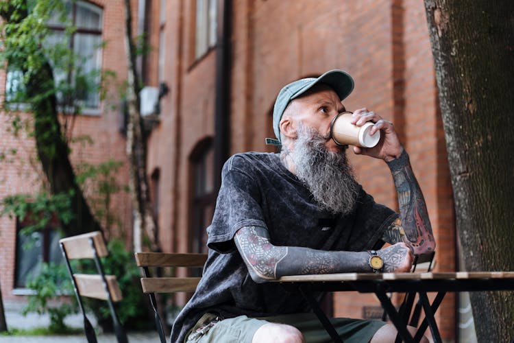 Bearded Man With Tattooed Hands Sitting At Table And Drinking Coffee At Outdoor Cafe