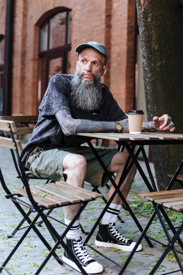 Adult Man In Cap On Head With Grey Beard Sitting At Coffee Table At Outdoor Cafe