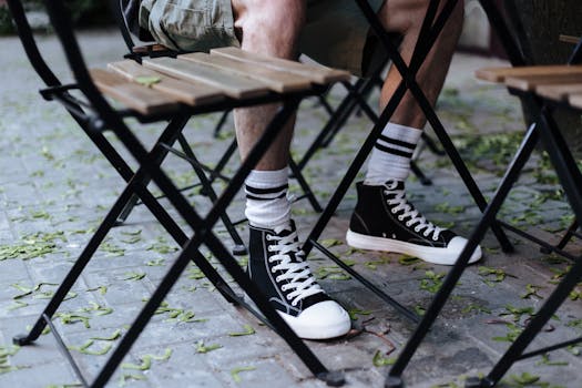 A close-up of a person in sneakers sitting at a cafe with scattered leaves on the pavement.