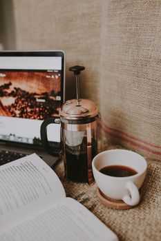 Inviting workspace scene with a cup of coffee, French press, laptop, and open book.