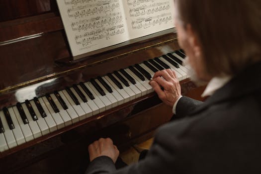 An elderly pianist plays a piano with sheet music, focusing on the keys in an indoor setting.
