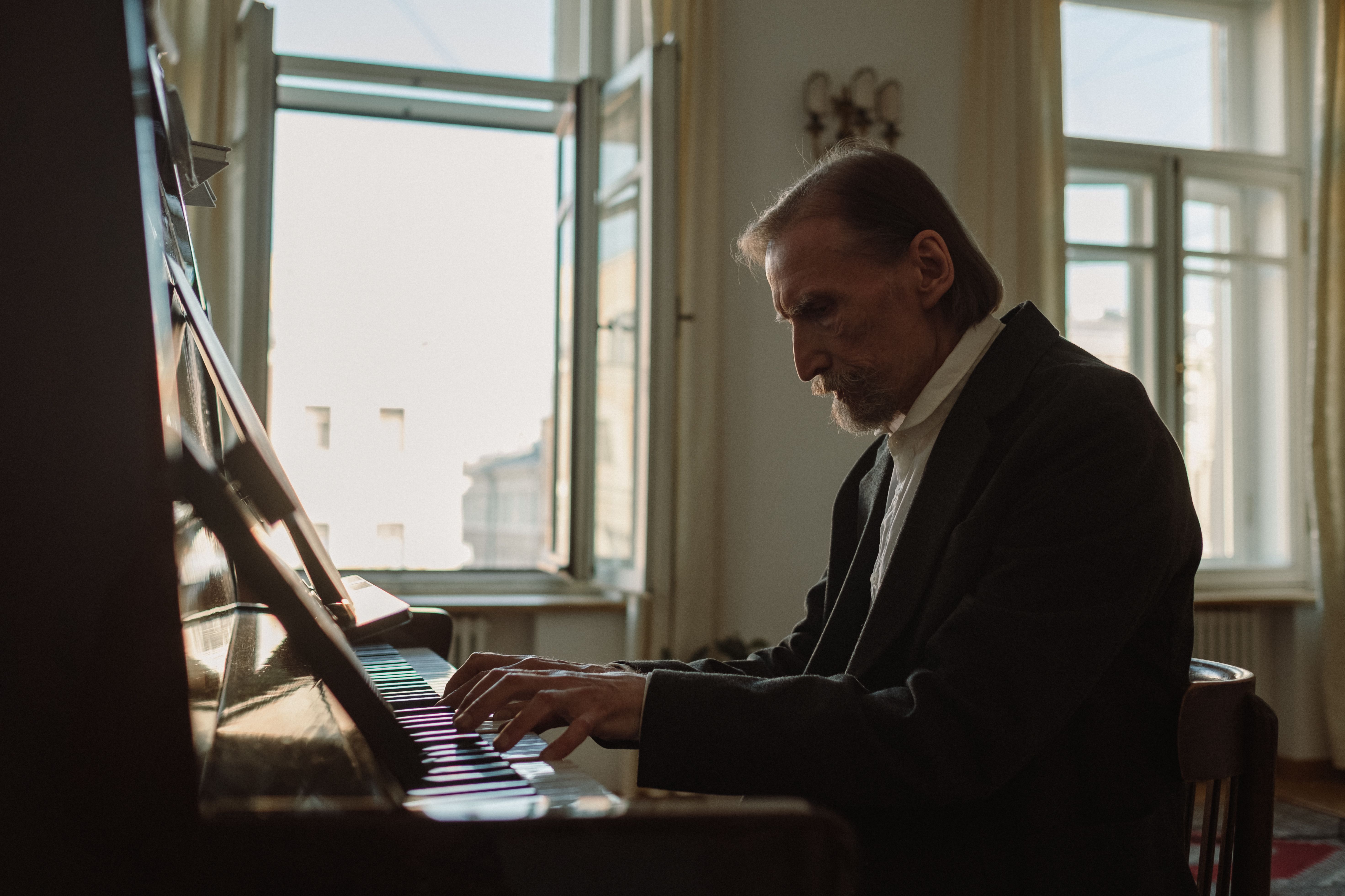 An elderly man thoughtfully playing the piano indoors in a sunlit room.