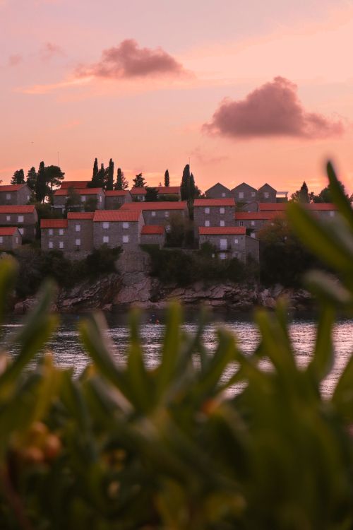 Free Serene view of a coastal village in Montenegro against a pastel sunset sky, framed by lush greenery. Stock Photo