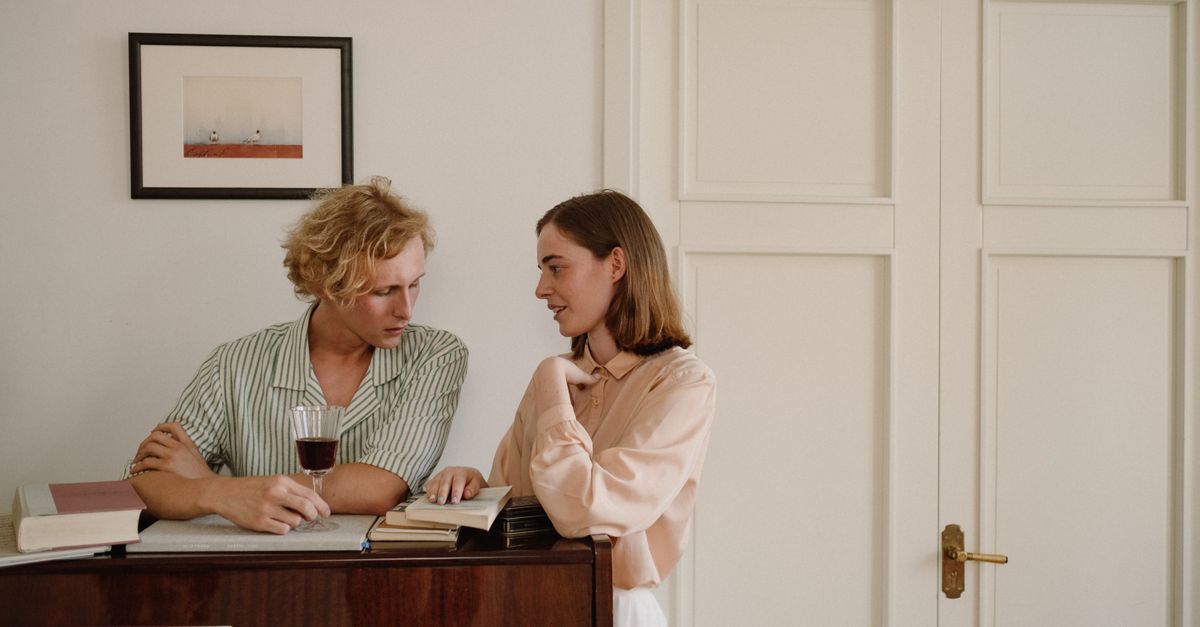 A couple engages in conversation by a piano, enjoying wine and books indoors.