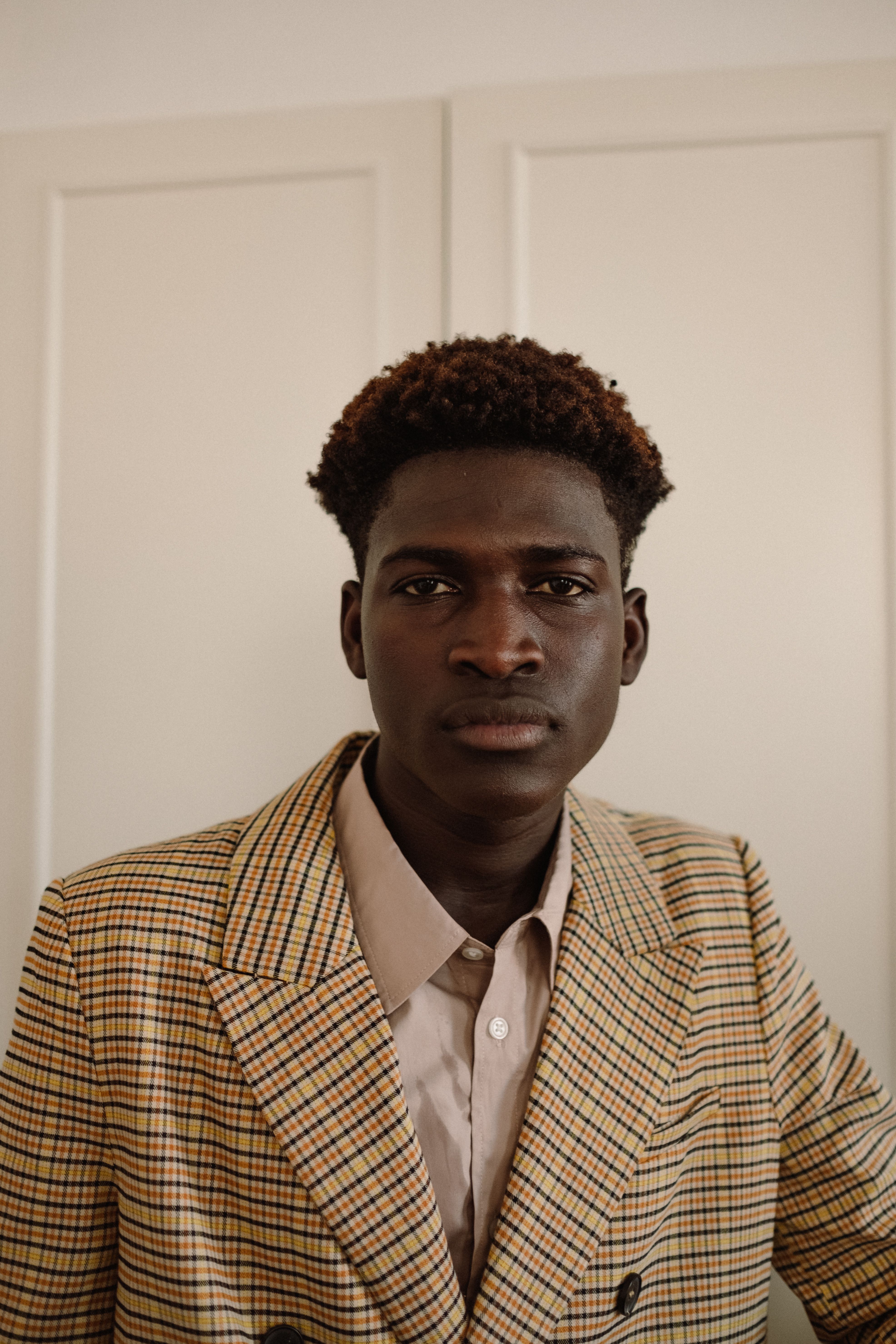 Stylish young man poses confidently in a plaid blazer against a neutral indoor backdrop.