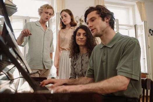 Group of young friends gathered around a piano, playing music indoors.
