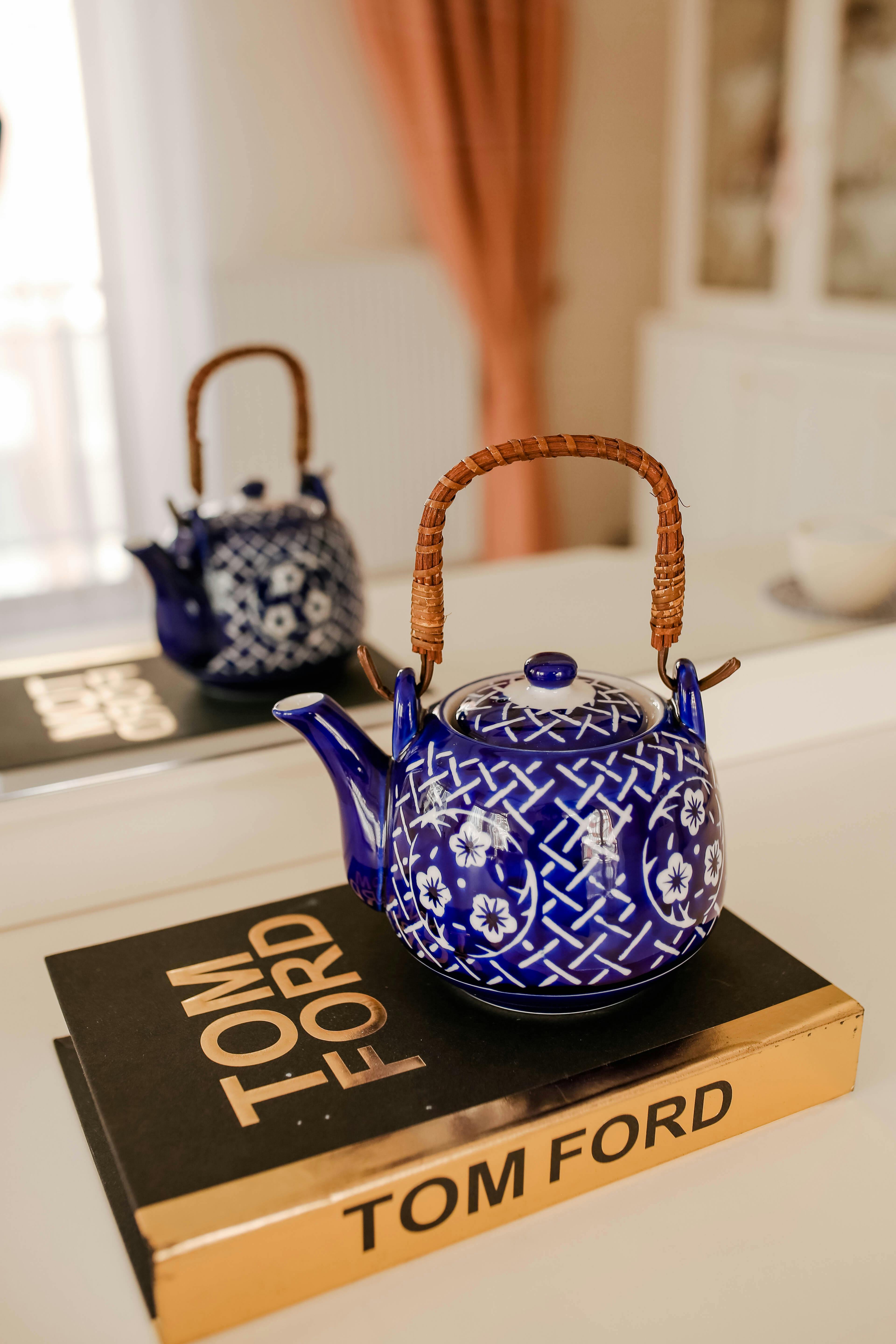 Blue floral teapot with wicker handle placed on Tom Ford book in stylish indoor setting.