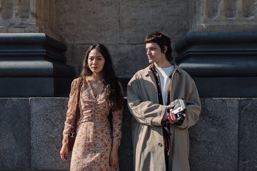 Two stylish individuals pose with an analog camera by a stone wall in daylight.
